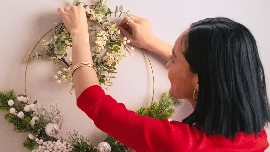 A woman with a dark bob and red top hangs a wreath on the wall using clear Command hooks