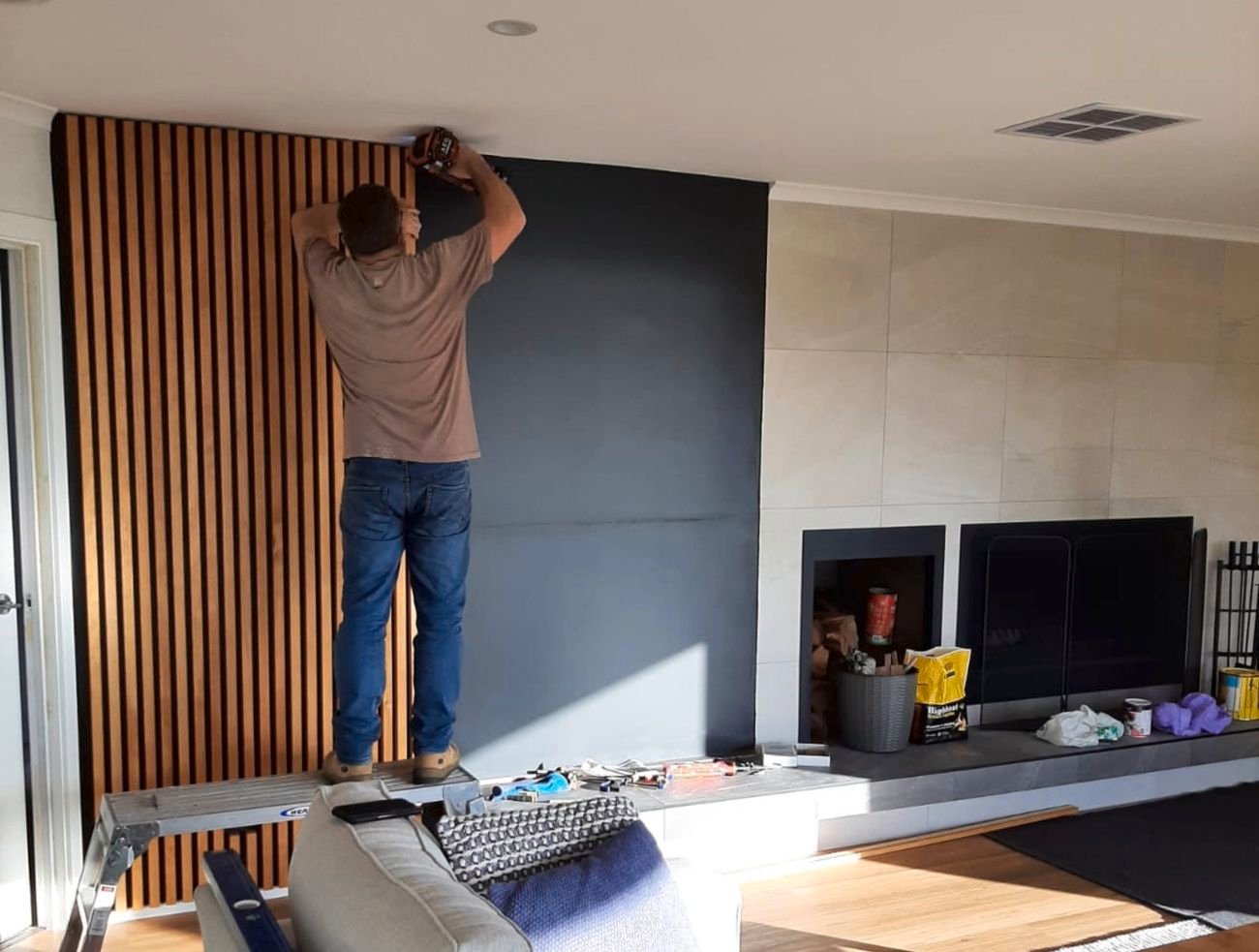 Man standing on ladder installing wooden slat wall in modern living room with fireplace and grey sofa.