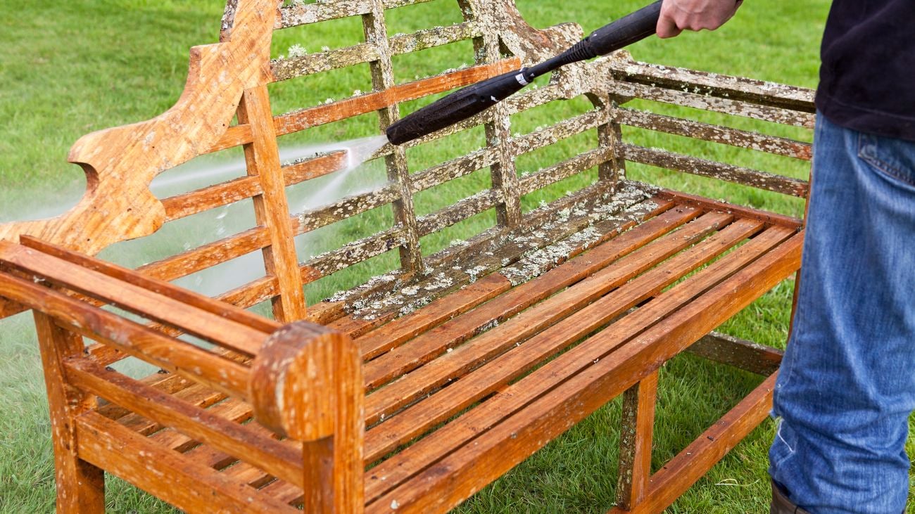 A person uses a pressure washer to clean and restore a teak wood Lutyens style bench.