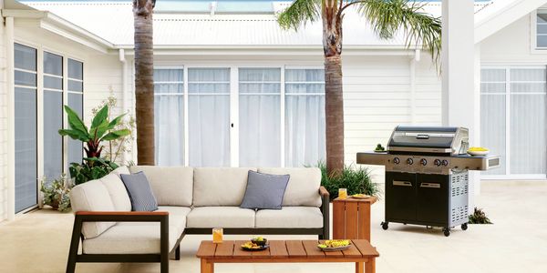 Covered outdoor entertaining area featuring a cushioned sofa, timber coffee table, barbecue and palm trees.