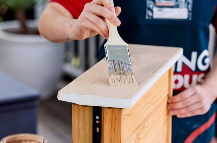 Person applying paint to the front of a bedside table drawer with a paintbrush.