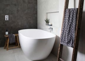 Grey and black tiled bathroom with white bathtub and towel hanging on rustic ladder.