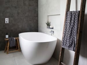 Grey and black tiled bathroom with white bathtub and towel hanging on rustic ladder.