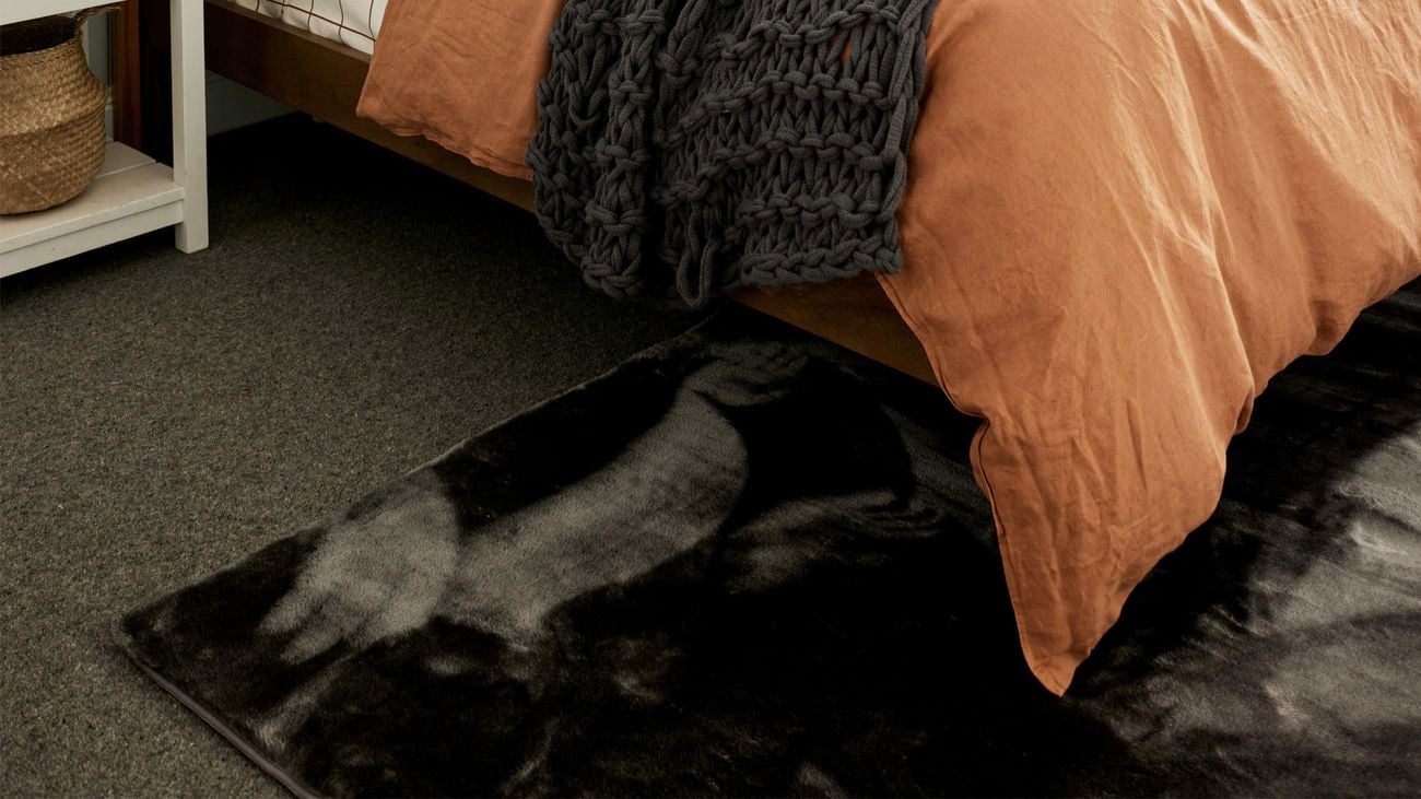 Corner shot of a bed with orange bedspread on top of a grey velvet rug.