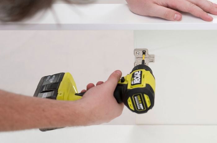  A person uses a drill to secure a hinge plate inside a cabinet beneath a shelf.