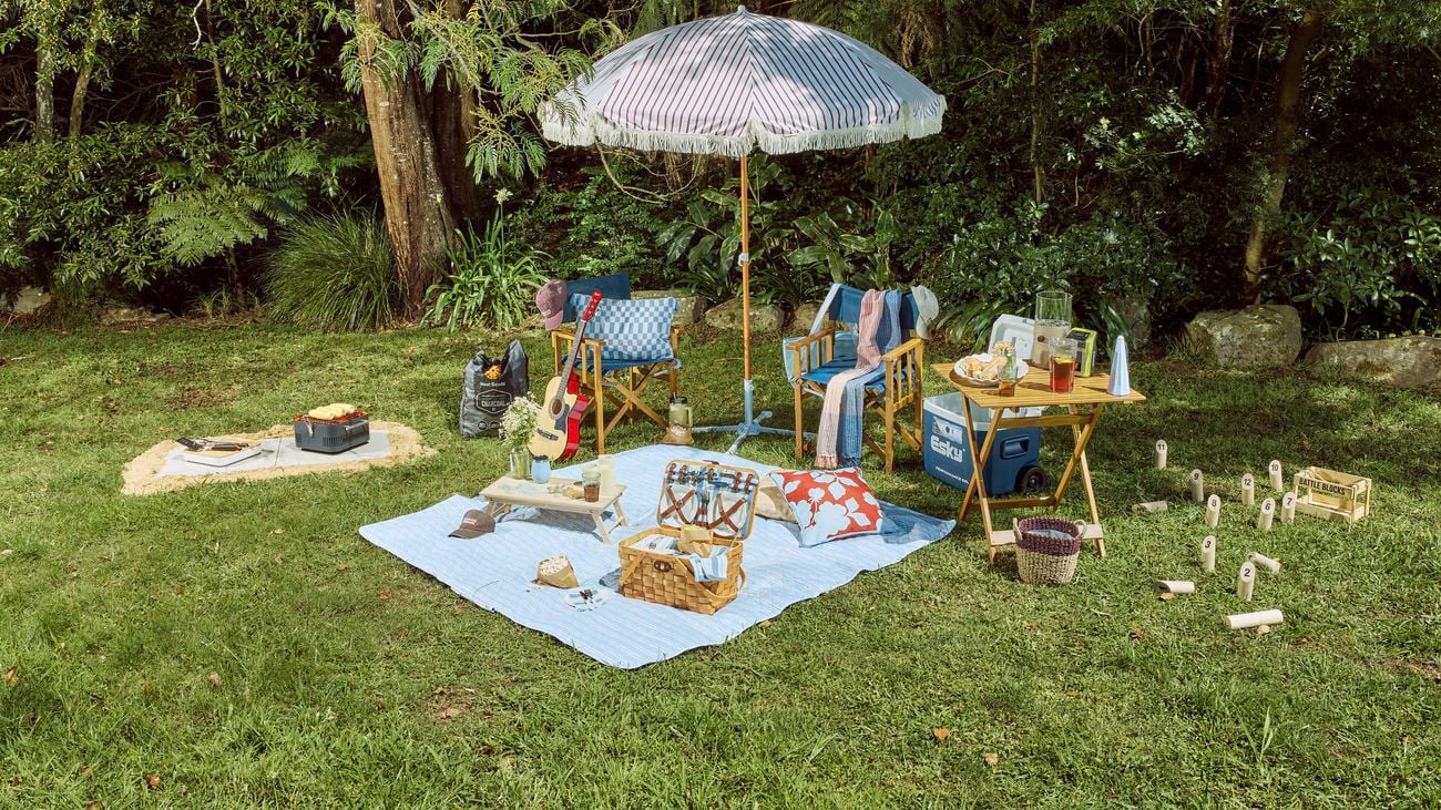 A styled outdoor picnic setup on a grassy lawn, featuring a blanket with baskets, drinks, and snacks, surrounded by wooden chairs, a cooler, and lawn games, all shaded by a large umbrella and set against a backdrop of trees.