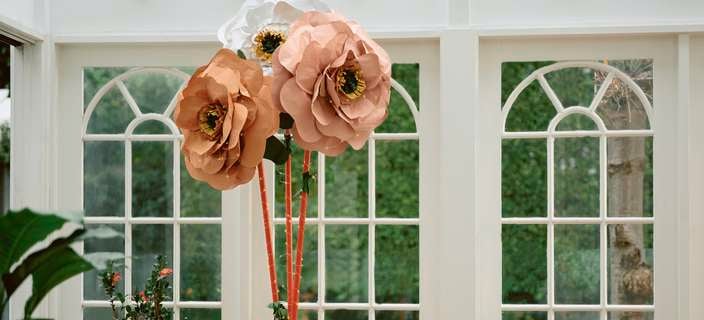 Three giant paper flowers in peach and white colours, surrounded by potted plants in a sunlit room with large windows.