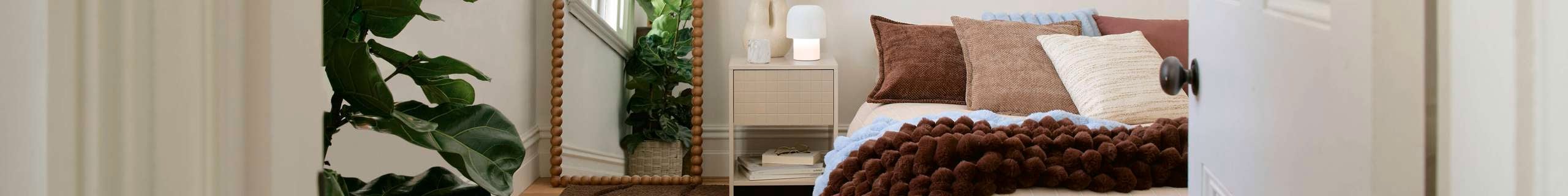 View into a bright, minimal bedroom with a low bed dressed in neutral bedding and brown throw, a brown rug, a tall floor mirror, a leafy indoor plant, and a white bedside table near tall window
