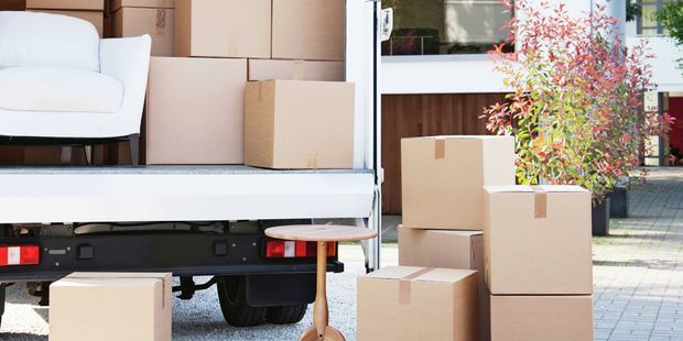  An open moving truck outside a modern house, filled with cardboard boxes and a white couch. Boxes and a small wooden table sit on the driveway.