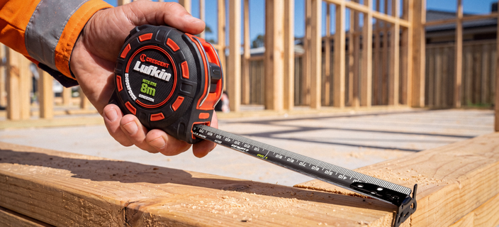 Close-up of a Crescent Lufkin Nite Eye tape measure extended across a timber beam while a worker marks measurements with a pencil.