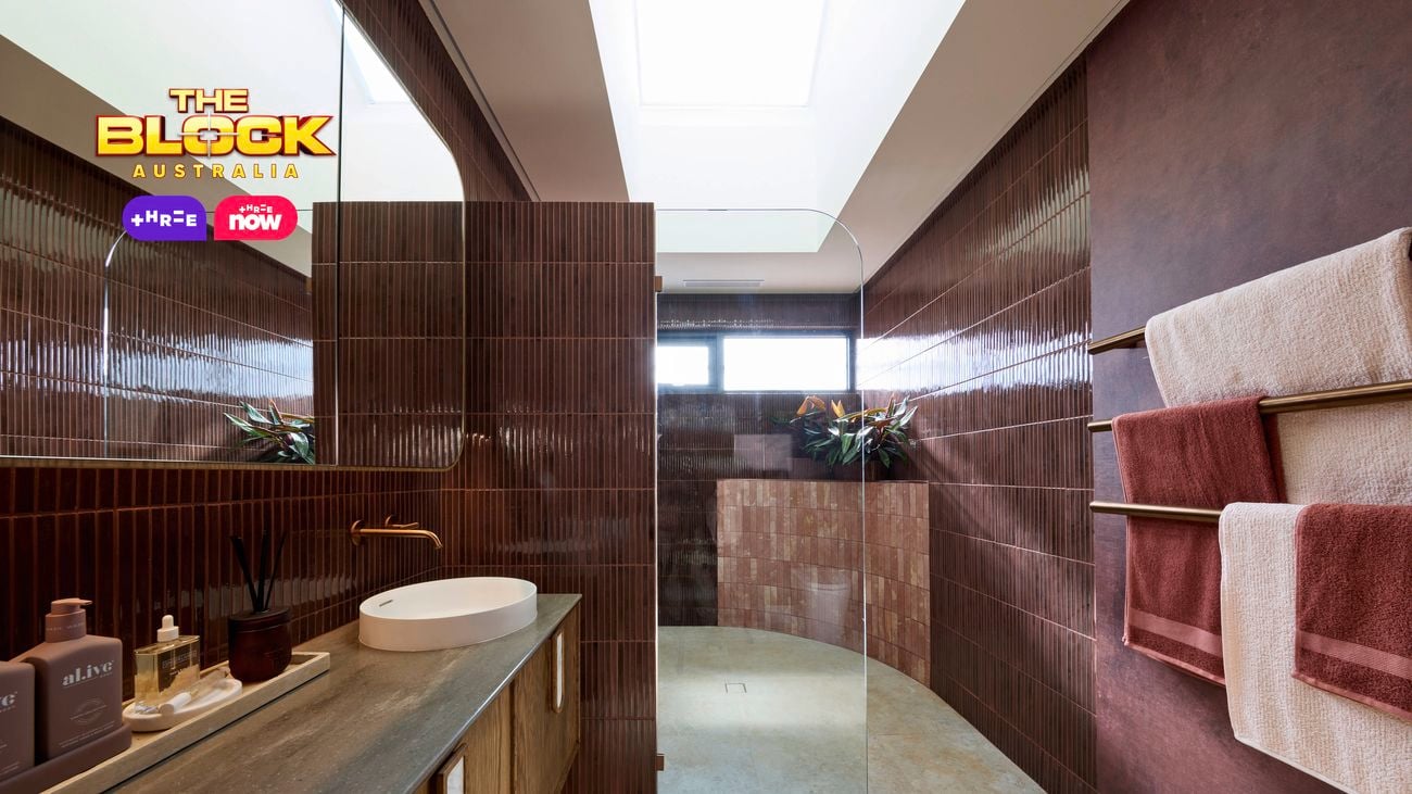 Modern bathroom with wooden vanity, oval sink, brass fixtures, glass shower, maroon tiles, towel racks, and skylight.