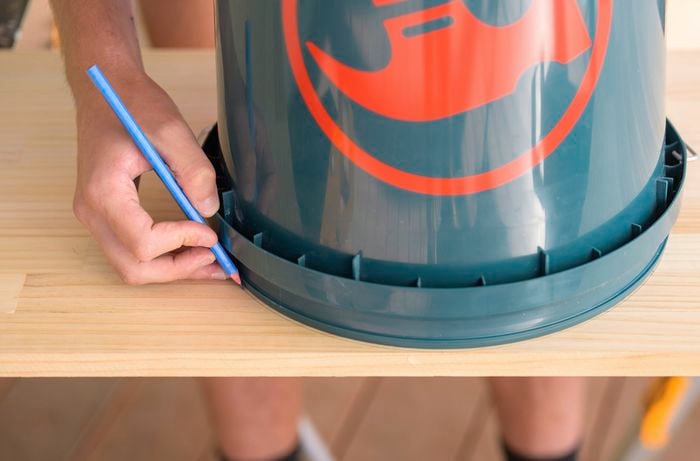 A person is using a bucket as a guide to sketch out wide, flowing half circles along one edge on a wooden board with a pencil.