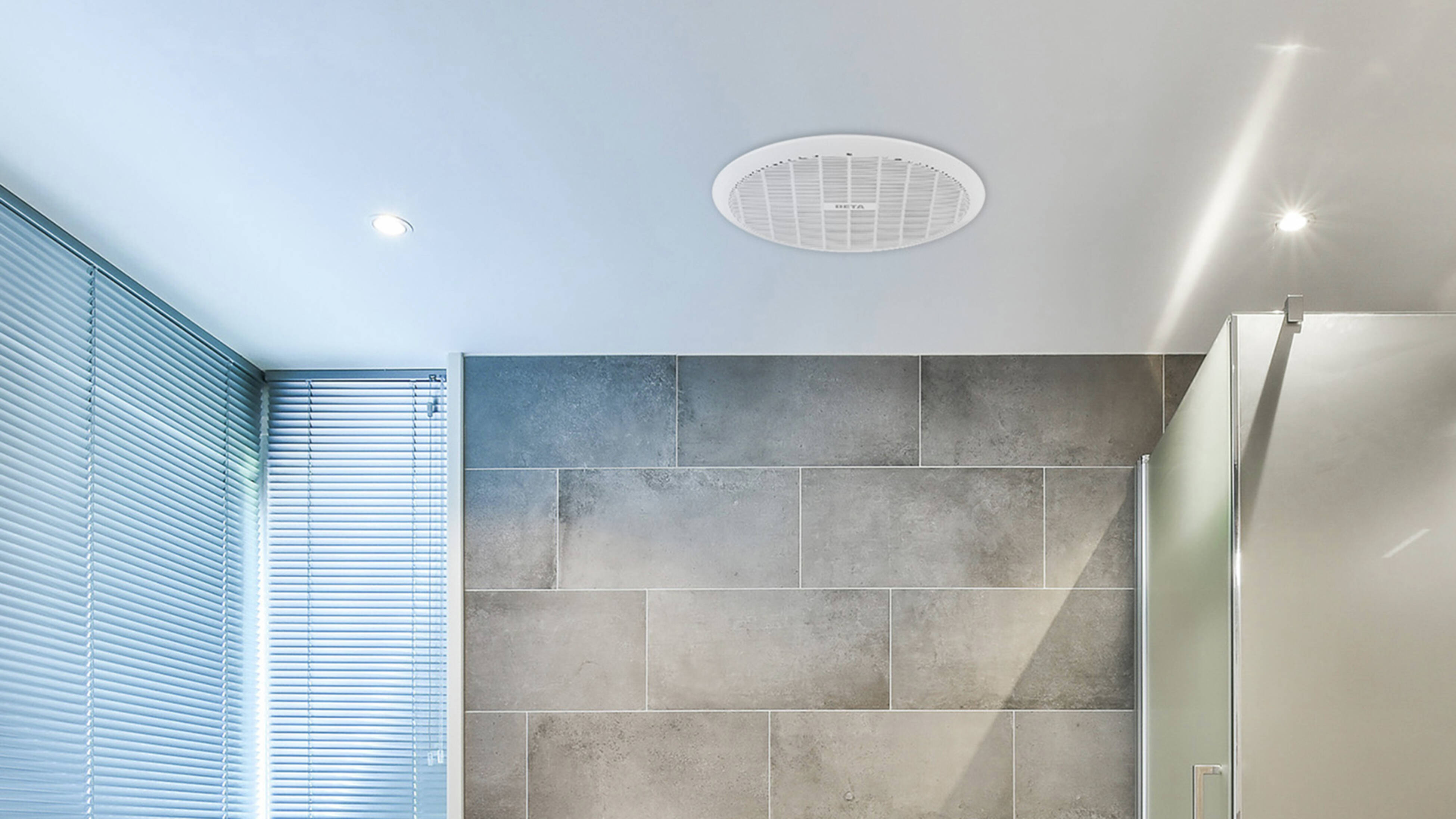  A modern bathroom with grey tiled walls and floor, a white freestanding bathtub, a window with blinds, a glass shower, and a ceiling exhaust fan.