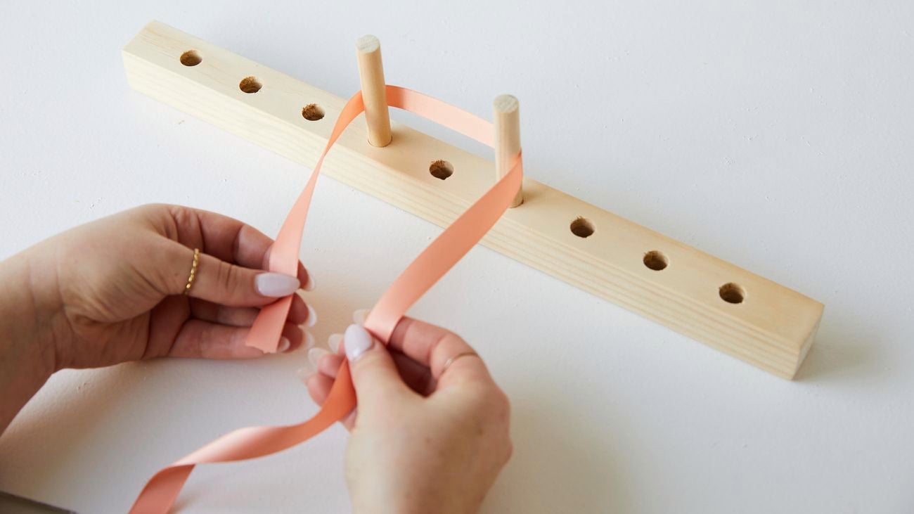 A person wraps a peach coloured ribbon around a timber bow maker.
