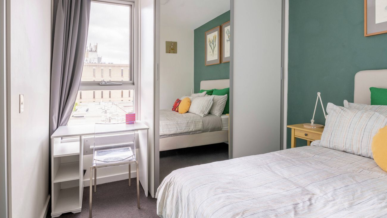 Bedroom with a green feature wall, a bed, a window with a desk underneath, and a large mirrored wardrobe reflecting the room.