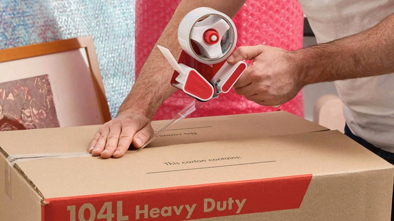 Close up of hands using a red tape dispenser to seal a large cardboard box, with bubble wrap and framed items in the background