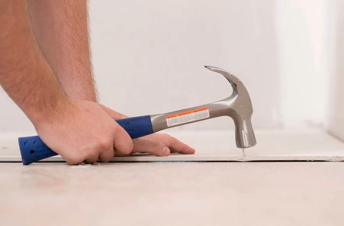 A person is using a hammer to secure a nail into an underlay sheet near the corner of a room.