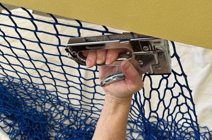 A hand uses a staple gun to attach a blue net to a yellow-painted timber surface.