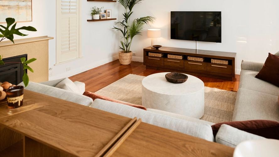 Light-filled living room with wooden floors, a wall-mounted TV above a low timber cabinet with baskets, a round coffee table on a rug, neutral sofas, indoor plants, and minimalist wall shelves.