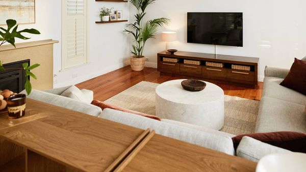 Light-filled living room with wooden floors, a wall-mounted TV above a low timber cabinet with baskets, a round coffee table on a rug, neutral sofas, indoor plants, and minimalist wall shelves.