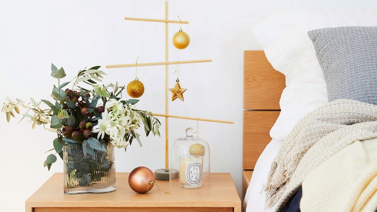 A wooden bedside table with a vase of flowers, a candle and ornaments next to a bed with white sheets and layered blankets, with a black pendant light above.