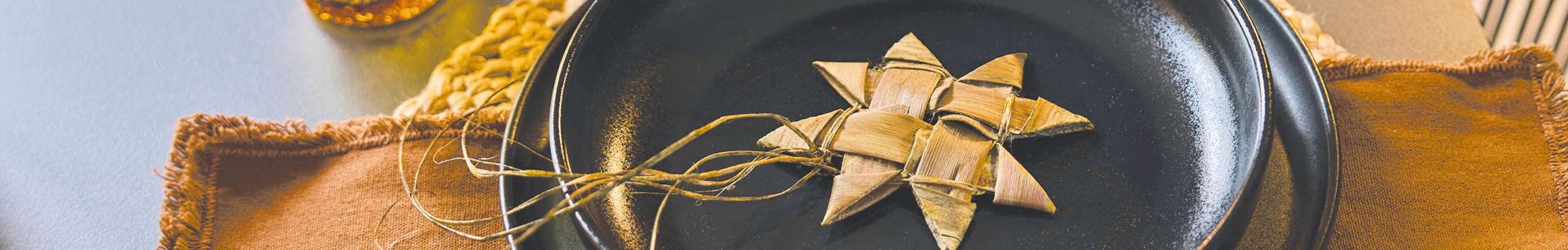 Close-up of a woven star decoration made from natural fibres placed on a black plate, styled with warm-toned table setting and textured fabrics.