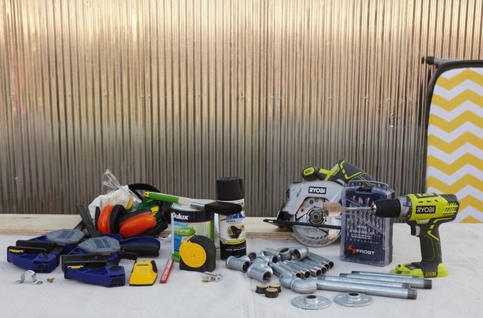 Tools and equipment for the job, including a power drill, lengths and joints of metallic pipe, spray paint, a can of paint, paintbrush, a stud finder, tape measure, wall brackets, clamps and more