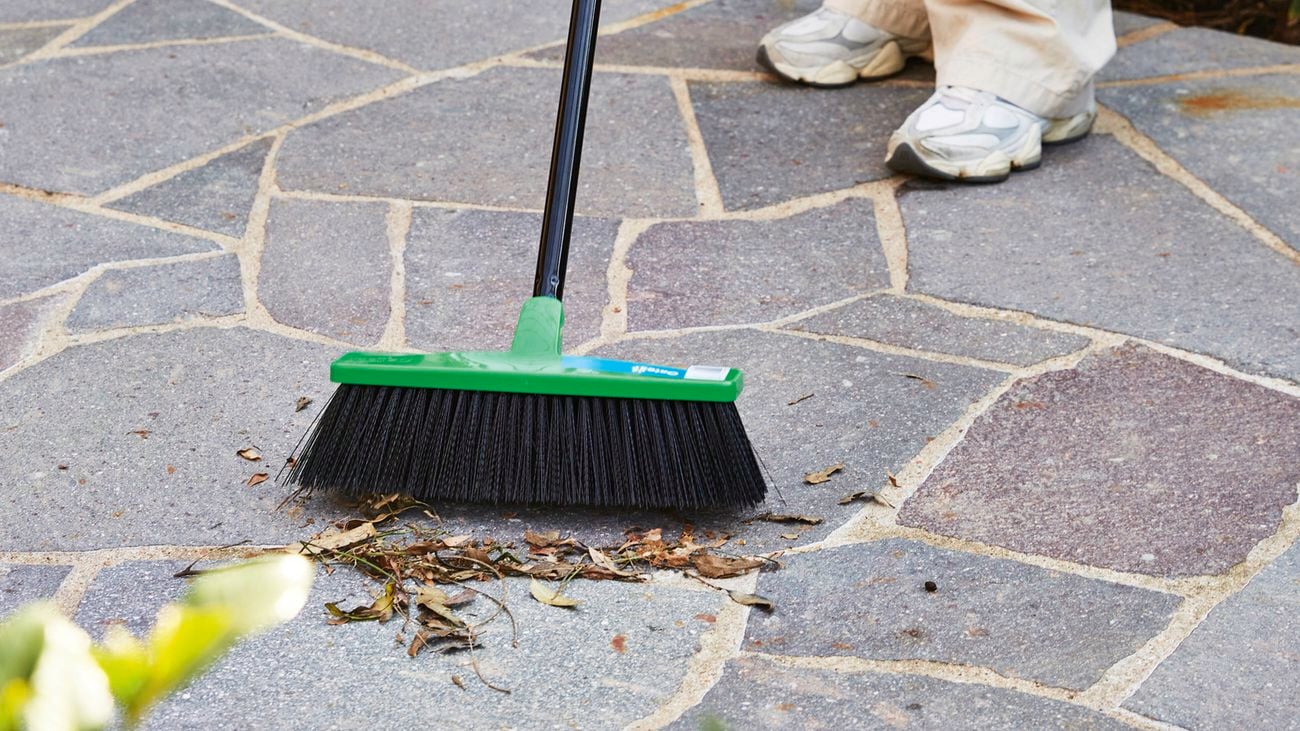 A person wearing white sneakers and light-coloured pants sweeps leaves and debris from a stone patio with a green and black broom