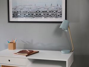 Painted metal desk lamp sits on a desk in front of a grey wall.