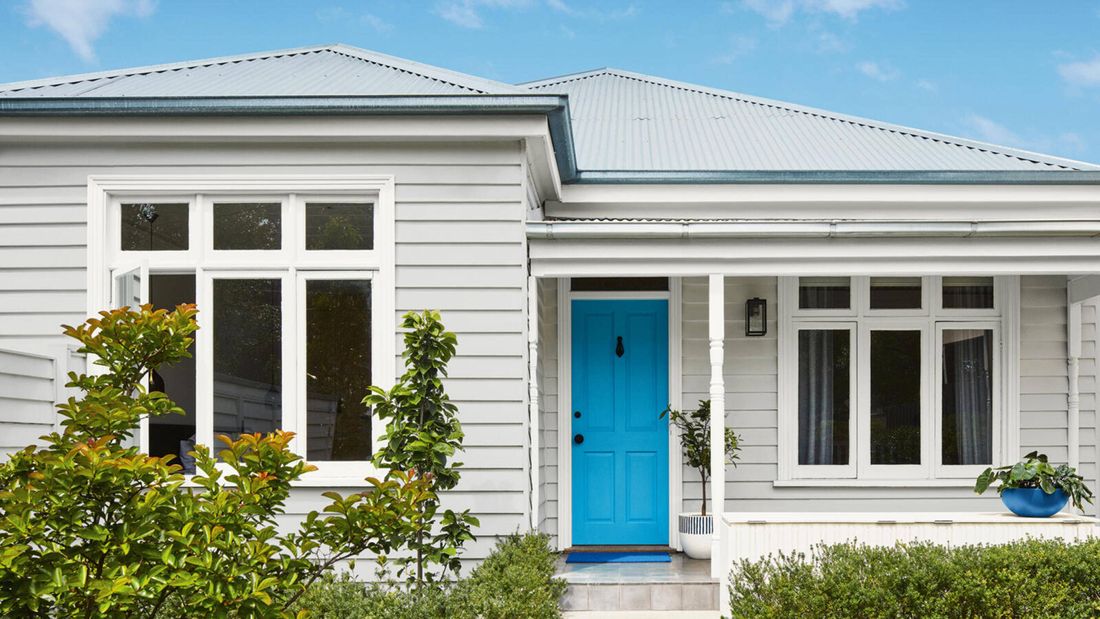 A white house with a bright blue front door, surrounded by green shrubs and a grey-tiled porch