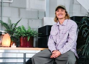 Person sitting indoors beside potted plants and a salt lamp, wearing a cap and patterned jacket.