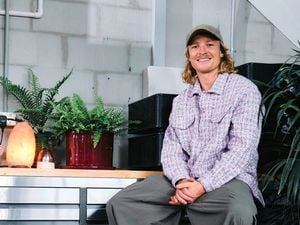Person sitting indoors beside potted plants and a salt lamp, wearing a cap and patterned jacket.