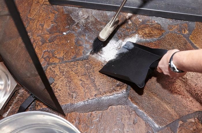 A person using a shovel and brush to sweep up ash in front of a wood heater