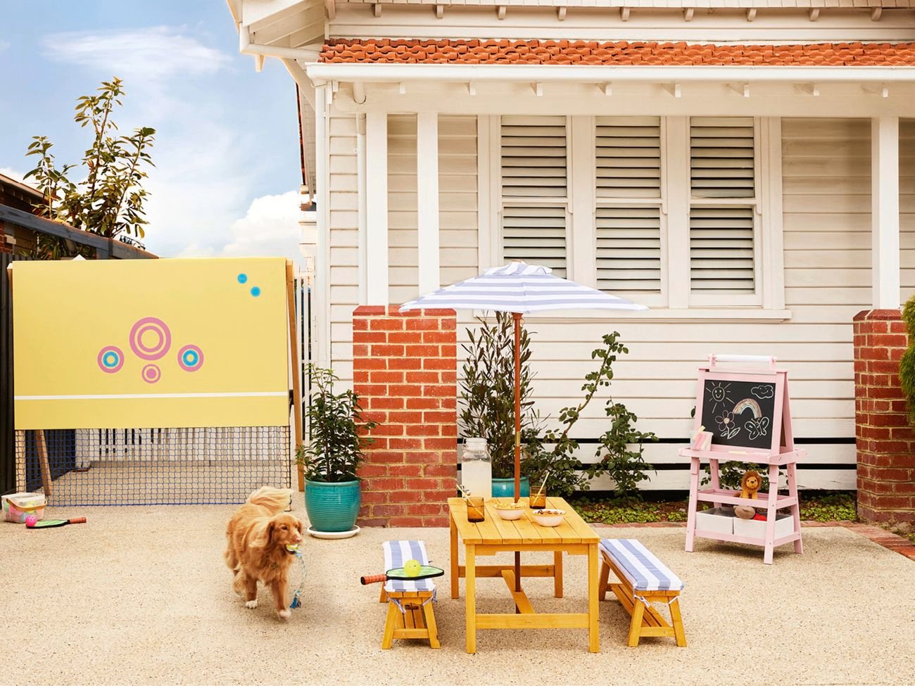 Dog carrying a toy walks across a paved front yard featuring a children’s tennis net, outdoor table with benches and umbrella, and a chalkboard easel in front of a weatherboard house.