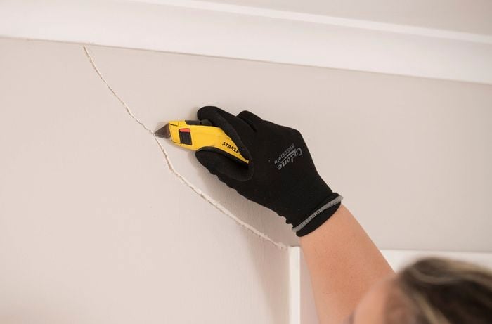 A person wearing gloves is using a utility knife to widen a crack in a wall, preparing it for repair