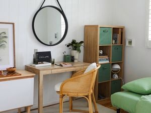 A bright guest room with white walls, a wooden desk and cane chair with a small plant and welcome chalkboard. To the right is a cube shelf with books and boxes, and a single bed with green bedding.