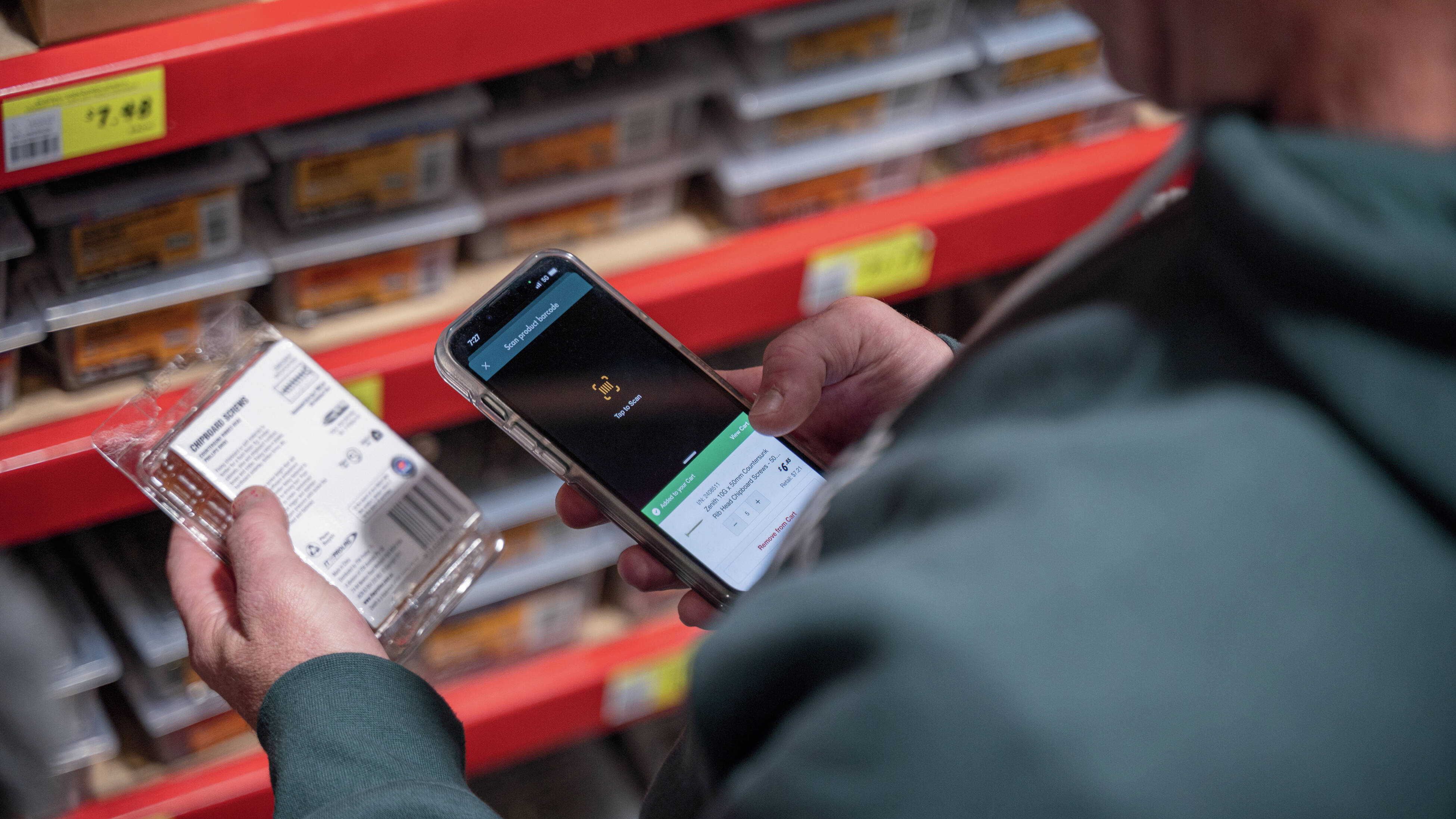 Close up of a tradesperson using a smartphone to scan the barcode of a pack of screws in a hardware store aisle.]