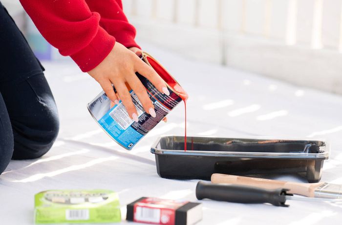 Pouring red exterior paint into a paint tray beside painting tools.