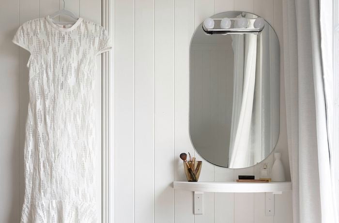  A makeup vanity table attached to a wall clad in white VJ panels alongside a hanging white dress