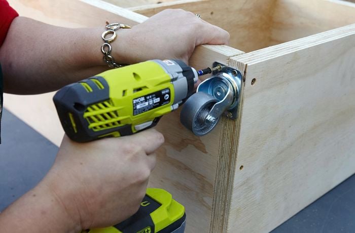 A person attaching a castor wheel to a plywood panel using a cordless drill