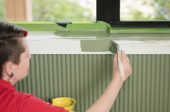 A woman, wearing a red T-shirt, is using a paintbrush to apply green paint to the architraves