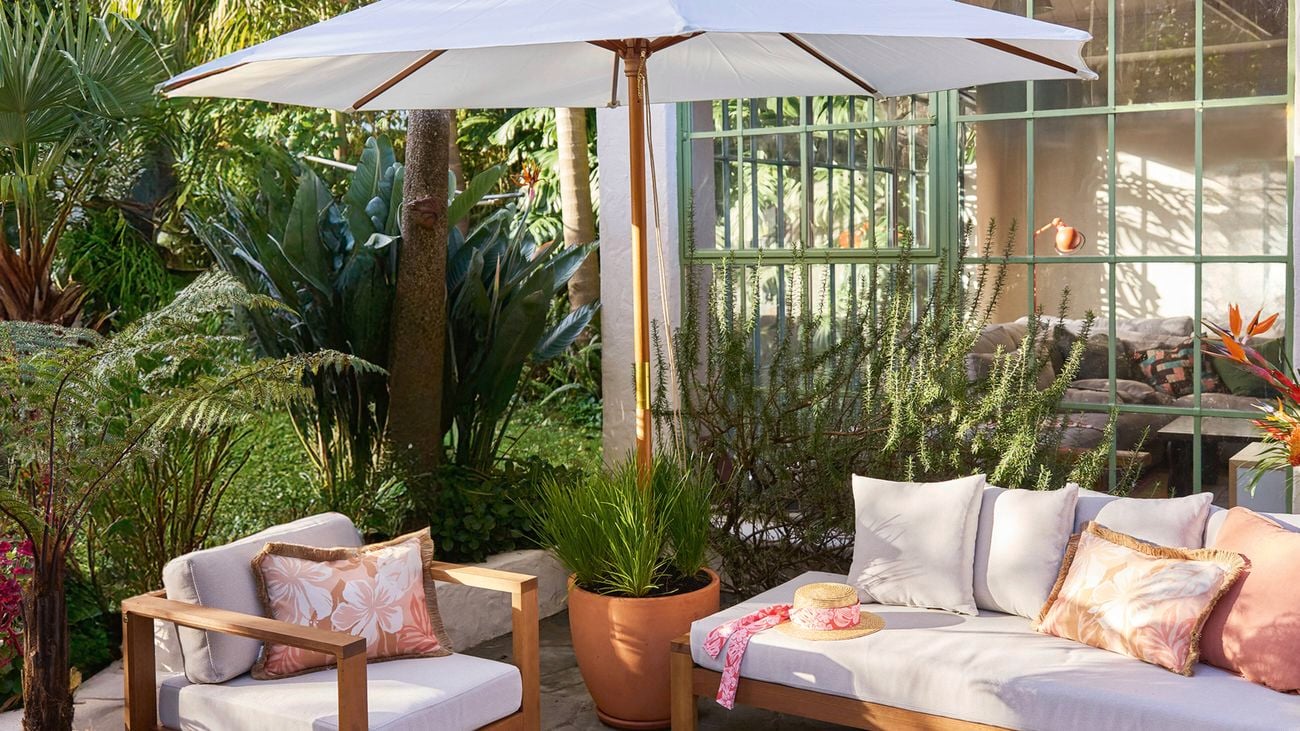  An outdoor garden design setting with a large umbrella offering shade, a white and timber lounge set and tropical greenery.
