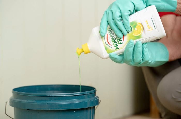 A person wearing gloves is preparing a cleaning solution by pouring dishwashing liquid into a bucket.