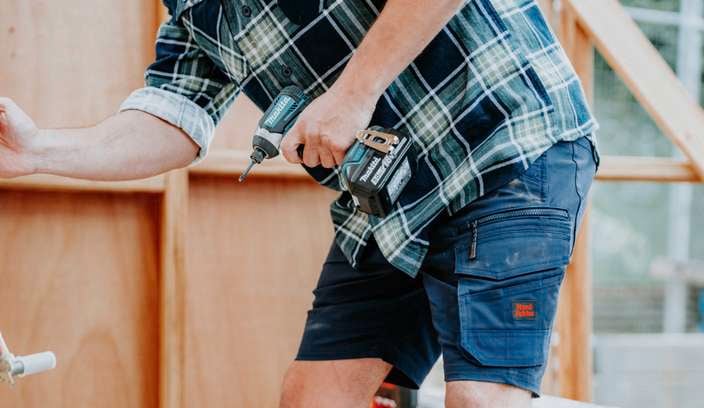 A person at a construction site with their hand touching a hose connector and the other hand holding a cordless drill.