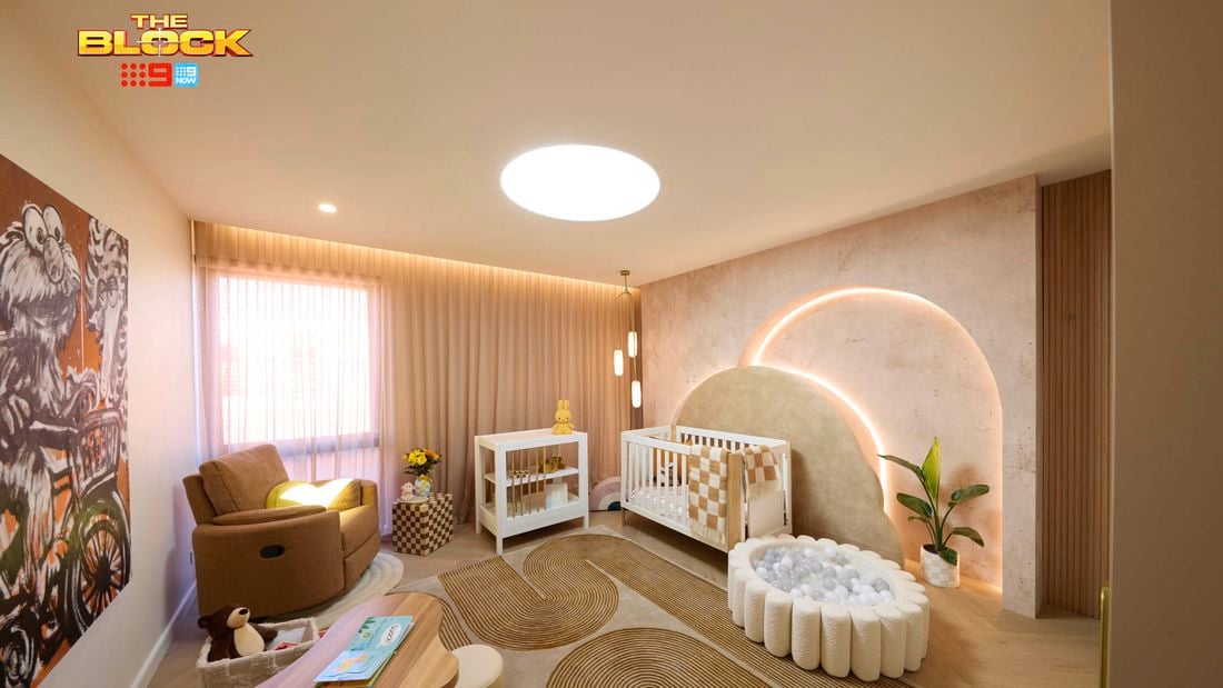 Neutral baby bedroom with brown and tan colours. Showcasing a cot and change table in front of an arched feature wall with feature lighting. There is a large chair in the corner and a play area on an arched pattern rug.