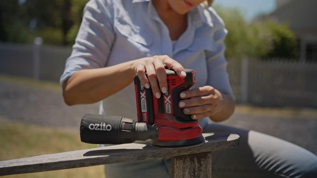 A pair of hands holding a random orbital sander to smooth back a weathered outdoor timber chair in a garden