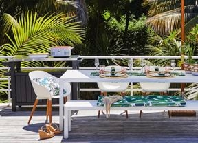 White outdoor table setting with glasses, cushions and pot plants.