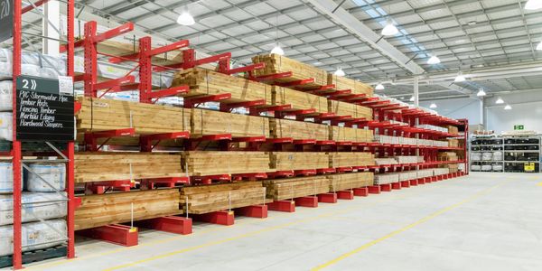 Long rows of timber beams and posts stored on red cantilever racking inside a spacious warehouse. The well-lit aisle has clear floor markings and signage identifying timber, fencing, edging and hardwood products.