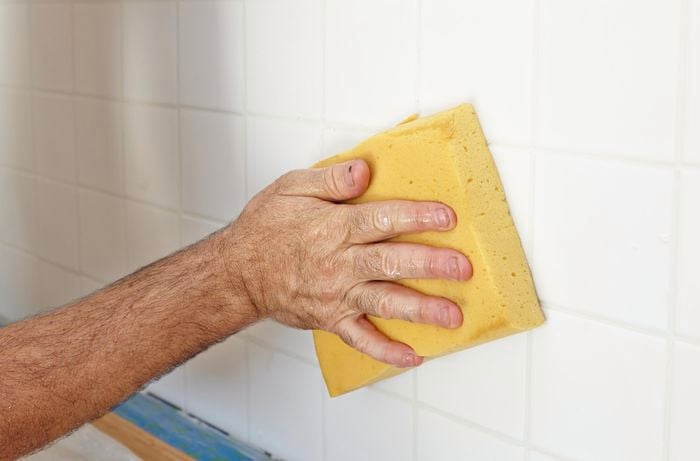 Person wiping freshly tiled splashback with wet sponge.