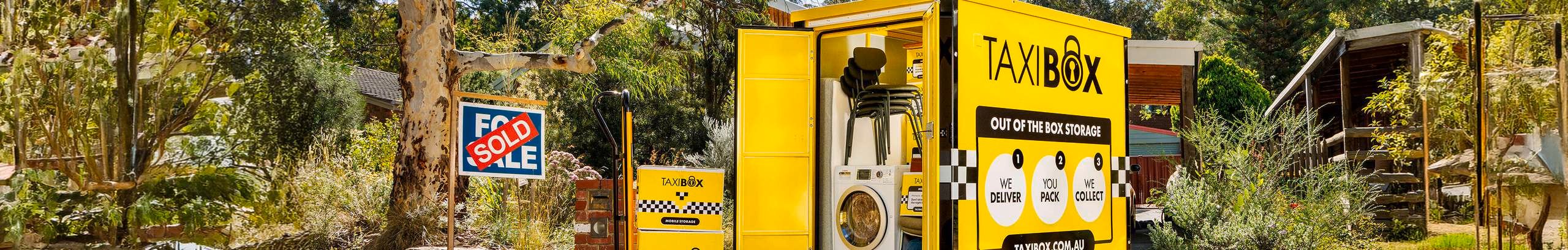 Yellow TAXIBOX storage unit parked outside a home, filled with household items like chairs and a washing machine, with a sold sign and garden setting in the background.